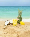A refreshing tropical drink beside a pineapple and broken coconuts on a sandy beach with the ocean in the background.