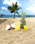 Tropical beach scene with a pineapple, split coconut, a glass of lemonade, and a lemon on the sand with palm trees and the ocean in the background.