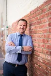 Man in a dress shirt and tie smiling with arms crossed standing against a brick wall.