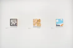 Three pieces of art displayed on a white wall in an exhibition, each with a descriptive label underneath.