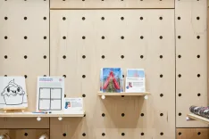 Interior of a store with wooden pegboard walls displaying greeting cards, an art experiment sign, and a canvas with a cartoonish ice cream drawing.
