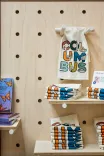 A wooden pegboard shelf displaying books, colorful tea towels with designs, and a white tote bag with "COLUMBUS" illustration hanging on a hook.