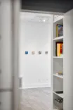 View through a doorway into a bright gallery room with a wooden floor and white walls displaying small artworks, adjacent to a bookshelf with colorful books.