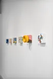 Contemporary art installation featuring a series of colorful abstract paintings and a yellow ceramic bowl displayed on white shelves against a white wall.