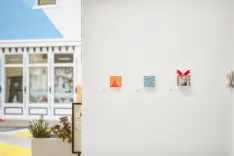 Contemporary art exhibition with a focused view of three square artworks on a white wall, with a blurred background showing gallery windows and a "We Can Do It" poster with a red ribbon.