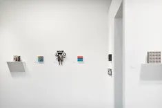 A modern art gallery interior with various small artworks mounted on a white wall and one artwork placed on a projecting shelf.