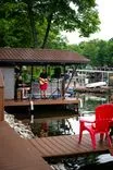 A person playing a guitar on a lakeside gazebo with red chairs in the foreground and docked boats in the background.