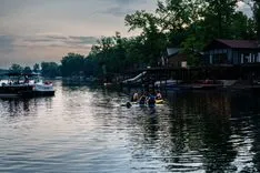 "Tranquil evening scene at a riverside with silhouettes of people on a small dock, houses along the shore, and boats on the water under a dusky sky."