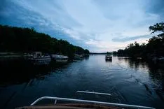 Twilight view from a boat on a serene river with forested banks and other boats docked along the shore.
