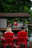 Two people sitting on red chairs by a river watching a musician perform on a small dock surrounded by greenery.