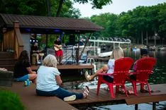 People sitting on a dock by a lake enjoying a live music performance at an outdoor venue.