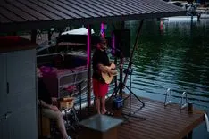 Musician playing guitar on a floating dock stage under a shelter at dusk.