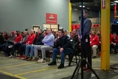 Group of people sitting on chairs in an industrial setting attentively listening to a presenter standing off-camera.
