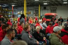 Group of people wearing red sitting in a factory setting with a vehicle in the background.