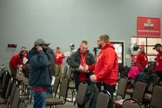 People in a room wearing warm clothing, some with red jackets, chatting and shaking hands, with folded chairs in the background and a red sign that reads "ENTRY."