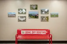 Red metal bench with "Bone Dry Roofing" logo in front of a beige wall displaying various framed pictures.