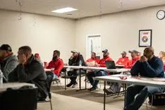 Group of people in red shirts seated at tables in a training session or meeting inside a room.
