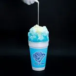 Condensed milk being poured on a blue raspberry snow cone in a branded cup against a black background.