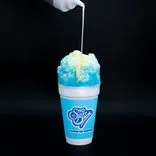 Condensed milk being poured on a blue raspberry shaved ice in a Sno Biz cup against a black background.
