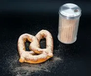 A cinnamon-sugar coated pretzel next to an overturned spice shaker on a black background.