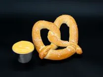 A freshly baked pretzel sprinkled with coarse salt next to a small container of melted cheese on a black background.