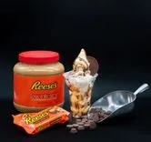 Jar of Reese's Peanut Butter Sauce, Reese's candy bars, and a sundae with peanut butter cups and chocolate chips on a black background.