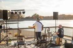 Two musicians performing on a lakeside deck during daytime with audio equipment around them.