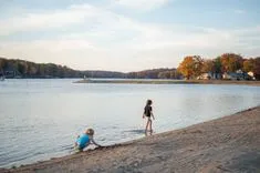 Two children playing by a calm lakeside with one standing in the water and the other sitting on the sand, during late afternoon.