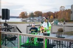 Two musicians performing outdoors on a deck overlooking a lake with autumn trees in the background.