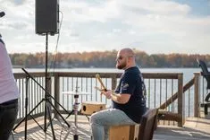 Musician with a guitar sitting on a chair by the lake, preparing for a performance with microphone stand and speaker in the foreground.