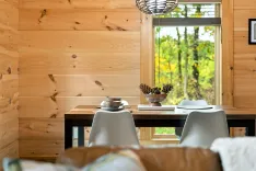 Cozy interior of a wooden cabin with a dining table set for two, ceramic tableware, decorative pine cones in a bowl, and a view of trees through a window.