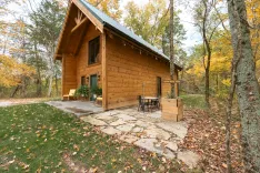 A cozy wooden cabin with a blue roof surrounded by autumn trees, featuring a front porch with chairs and a path leading to a sign that reads 'Cedar Ridge Trail'.