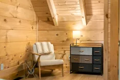 Cozy wooden cabin interior with a soft armchair, a fabric-covered chest of drawers, and a lamp on top, illuminated in warm light.