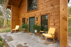 Exterior view of a wooden cabin with a concrete patio featuring two yellow chairs, potted ferns, and a welcoming atmosphere amidst autumn foliage.