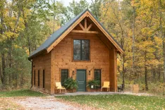 A cozy wooden cabin with a gabled roof and a front porch surrounded by autumn trees.
