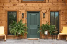 Cozy wooden cabin entrance featuring a green door flanked by large ferns and yellow chairs with modern outdoor lighting.