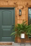 A welcoming home entrance featuring a green door with an arched design, surrounded by natural wood walls, a modern wall sconce, a lush fern plant in a white pot on a black wire stand, and a doormat with the text "Leave Your Worries Here".