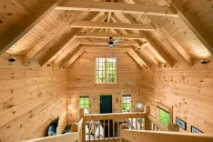 Interior view of a wooden cabin loft area with exposed beams, a ceiling fan, large windows, and a partial view of the staircase and furnishings.