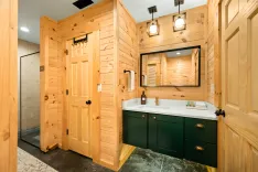 Rustic bathroom interior with wood-paneled walls, a glass-door shower, a vanity with green cabinets and white countertop, a large mirror, and industrial-style lighting fixtures.