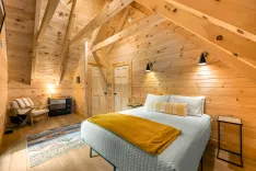 Cozy wooden attic bedroom with a double bed, armchair, dresser, colorful rug, and warm lighting.