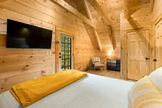 Cozy wooden cabin bedroom interior with a large bed, yellow throw blanket, mounted flat-screen TV, and a door leading to a balcony.