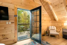 Cozy wooden cabin interior with open glass door leading to a balcony overlooking autumn trees.