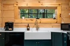 A stylish kitchen with wooden walls and a large window, featuring dark green cabinets, white countertops, a farmhouse sink, and a black coffee maker.