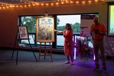 Two people presenting artwork at an indoor event, with string lights above and a window view of a waterfront in the background.