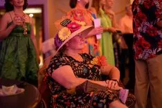 Woman wearing a festive hat and holding a fan while sitting at a social event with blurred figures in the background.