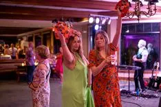 Two women dancing holding flower bouquets at a festive event with a live band in the background.
