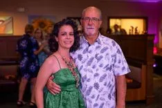 A woman in a green dress smiling alongside an older man in a patterned shirt, with blurred figures in the background at what appears to be a social event.
