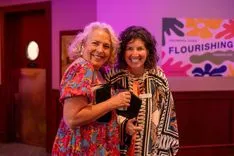 Two smiling women posing for a photo at an event with a colorful background featuring the word 'FLOURISHING'.